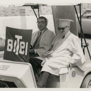 Two members of the Class of 1918 in graduation-style reunion gowns in golf cart at class reunion (Trinity College, Hartford Connecticut), 1968